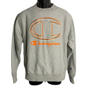 Puede incluir: Una sudadera gris Champion con un gran logotipo "C" de color naranja y negro con contorno y la palabra "Champion" en naranja debajo del logotipo.