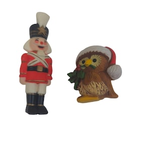 May include: A red and white toy soldier figurine with a black hat and a red and white uniform. Next to it is a brown owl figurine wearing a red and white Santa hat and holding a sprig of holly.
