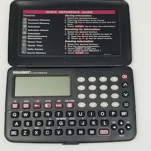 Könnte beinhalten: Ein schwarzer digitaler Organizer von Rolodex Electronics mit offenem Deckel. Das Gerät verfügt über eine QWERTZ-Tastatur, einen Taschenrechner und einen kleinen Bildschirm. Der Deckel zeigt eine Kurzanleitung mit Funktionen und Anweisungen.