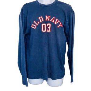 May include: A navy blue long-sleeve shirt with the text "OLD NAVY 03" in red and white lettering. The shirt has a ribbed texture and a crew neck. The shirt is displayed on a white mannequin.