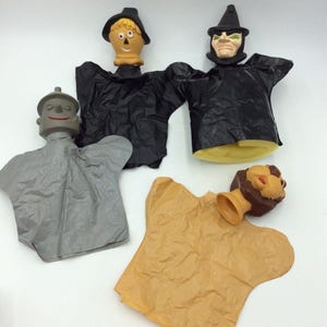 May include: Four vintage hand puppets featuring characters from The Wizard of Oz. The puppets include the Scarecrow, the Wicked Witch, the Tin Man, and the Cowardly Lion. Each puppet has a detailed head and a fabric body.