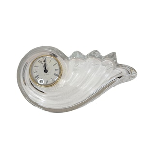 Puede incluir: Un reloj de cuarzo de cristal transparente con un bisel dorado y números romanos. El reloj está dentro de una base decorativa de cristal en forma de ola. Las manecillas indican la hora.