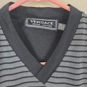 May include: Black and white striped V-neck shirt with a black collar. The shirt features the "Versace Jeans Couture" label in the center. The size is XL.