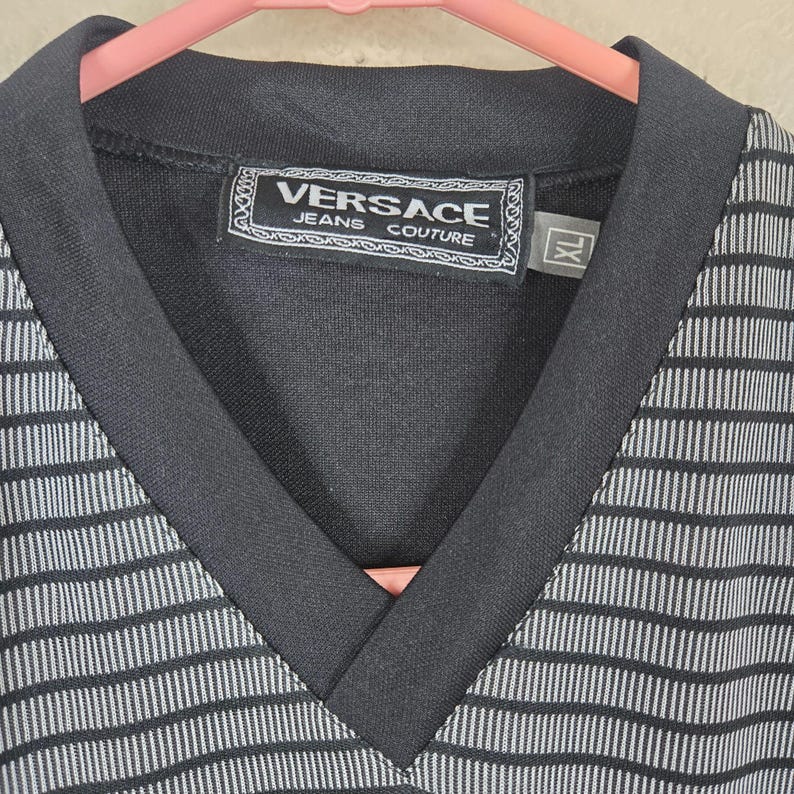 May include: Black and white striped V-neck shirt with a black collar. The shirt features the "Versace Jeans Couture" label in the center. The size is XL.