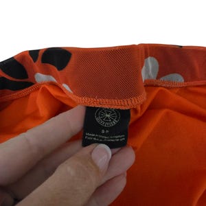 Pu&ograve; includere: Primo piano di un capo d'abbigliamento arancione con motivi bianchi e neri. Il capo ha un'etichetta nera con la scritta "OUTFITTERS" e il testo "Made in United Kingdom Fabriqu&eacute; au Royaume-Uni".