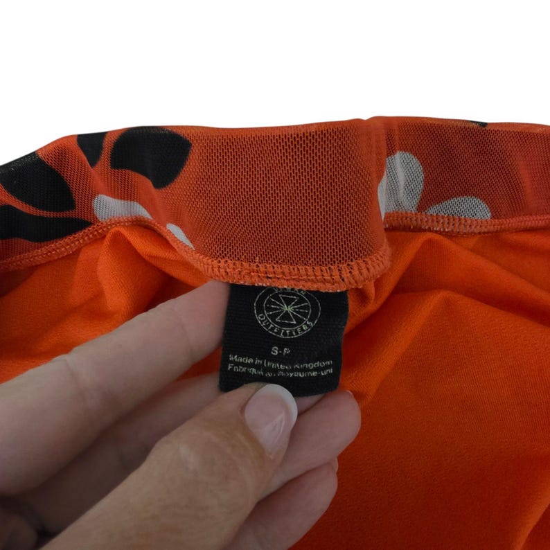 Pu&ograve; includere: Primo piano di un capo d'abbigliamento arancione con motivi bianchi e neri. Il capo ha un'etichetta nera con la scritta "OUTFITTERS" e il testo "Made in United Kingdom Fabriqu&eacute; au Royaume-Uni".