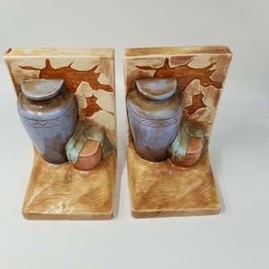 May include: Pair of ceramic bookends featuring a rustic design. Each bookend has a light blue vase, a small basket, and a draped cloth against a backdrop of a cracked wall. The color palette includes beige, brown, and blue tones.