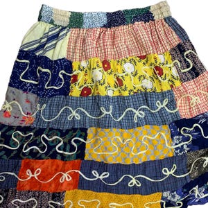 May include: A patchwork skirt featuring a variety of colorful fabric patterns. The skirt has an elastic waistband and is composed of squares and rectangles of different colors and designs, including floral, plaid, and abstract patterns. White decorative stitching accents the seams.