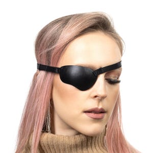 Puede incluir: Un parche de cuero negro con una correa ajustable se usa en la frente. El parche en el ojo lo usa una persona con cabello rosado y un suéter de cuello alto beige. El fondo es blanco.