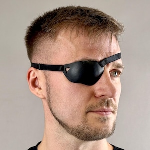 Peut inclure: Un cache-œil noir en cuir, couvrant un œil. Le cache-œil est maintenu en place par une fine sangle. La personne porte un haut noir. Le cache-œil est conçu pour s'adapter à la forme de l'œil.