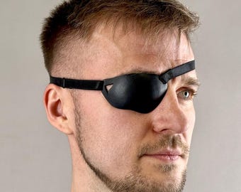 Op de afbeelding: Een zwarte leren ooglap, gedragen over één oog. De ooglap heeft een gebogen vorm om in het oog te passen en wordt op zijn plaats gehouden door een dunne band. De persoon draagt een zwart shirt.