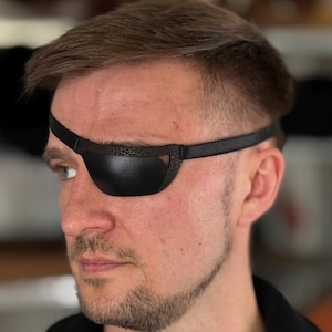 May include: A black leather eye patch with a strap worn across the forehead. The eye patch is designed to cover the left eye. The person in the image is looking to the left.