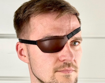Könnte beinhalten: Braune Leder-Augenklappe mit schwarzem Riemen, getragen über dem rechten Auge. Die Augenklappe ist so konzipiert, dass sie das Auge bedeckt und wird von einem schwarzen Riemen gehalten, der um den Kopf geht. Die Person im Bild trägt ein schwarzes Hemd.