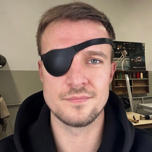 May include: Black leather eye patch with a strap, covering one eye. The eye patch is a solid black colour and designed to cover the eye completely. The strap is also black and appears to be made of leather.