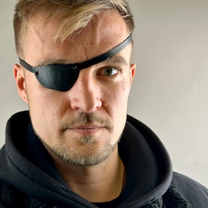 May include: A black eye patch with a strap worn across the forehead. The eye patch covers the left eye. The person is wearing a black hooded sweatshirt. The background is a solid light grey.