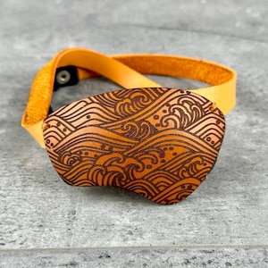 Può includere: Una benda sull'occhio in pelle marrone con un motivo a onde, attaccata a un cinturino in pelle arancione. La benda sull'occhio ha una forma ovale e presenta un motivo a onde inciso. Il cinturino ha una chiusura a bottone.