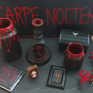 May include: A collection of gothic-themed decor items, including a black jar with red dripping accents, a wooden candlestick, a small chest with a bat emblem, and a framed picture. The background features the text "CARPE NOCTEM" in red.