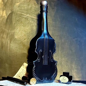 Può includere: Una bottiglia di vetro nero a forma di violino con un tappo dorato. La bottiglia è appoggiata su un pezzo di carta con una penna e un calamaio.