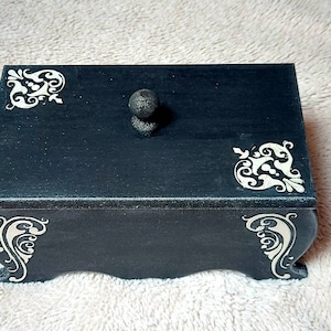 Op de afbeelding: Een zwarte houten doos met een decoratieve knop op de deksel. De doos heeft witte bloemmotieven op de deksel en de zijkanten.