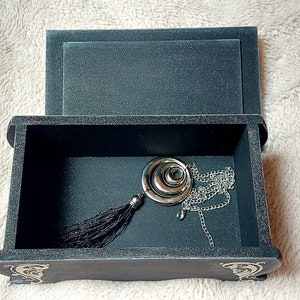 Op de afbeelding: Een zwarte houten doos met een scharnierend deksel, open om een ​​zilveren ketting met een ronde hanger en een zwarte kwast te onthullen. De doos heeft decoratieve details aan de voor- en zijkanten.