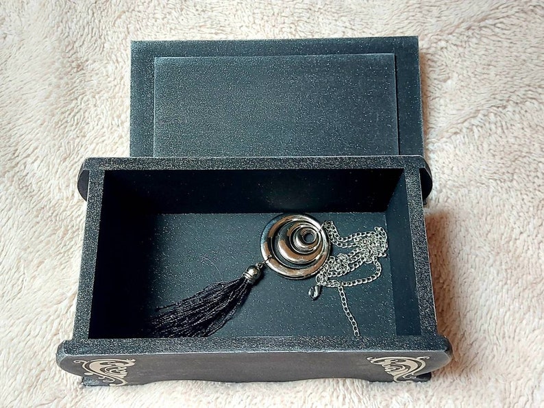 Op de afbeelding: Een zwarte houten doos met een scharnierend deksel, open om een ​​zilveren ketting met een ronde hanger en een zwarte kwast te onthullen. De doos heeft decoratieve details aan de voor- en zijkanten.
