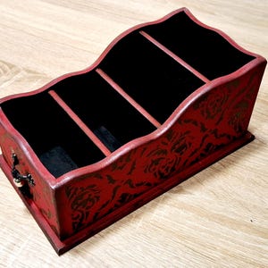 May include: A red wooden organizer with a black interior and four compartments. The exterior features a floral pattern and a small drawer with a decorative handle. The organizer sits on a wooden surface.