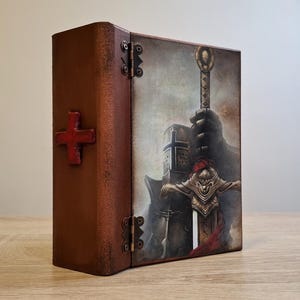 May include: A wooden box with a hinged lid, decorated with a red cross and a painted image of a knight holding a sword. The knight is wearing a helmet and armour.