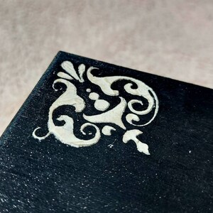 Op de afbeelding: Een zwarte doos met een wit decoratief ontwerp op de hoek. Het ontwerp is een gestileerd bloemmotief.