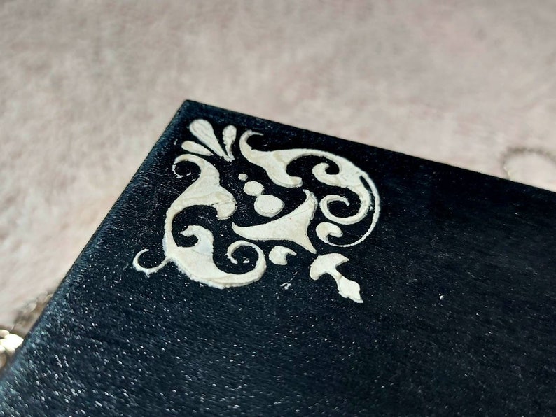 Op de afbeelding: Een zwarte doos met een wit decoratief ontwerp op de hoek. Het ontwerp is een gestileerd bloemmotief.