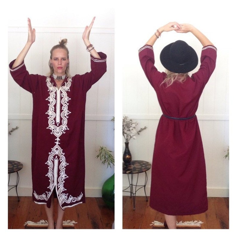 K&ouml;nnte beinhalten: Ein langes, burgunderfarbenes Kleid mit wei&szlig;en Stickereien. Das Kleid hat lange &Auml;rmel und einen V-Ausschnitt. Das Model tr&auml;gt eine silberne Halskette und einen schwarzen Hut.