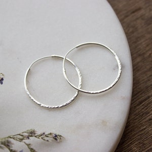 May include: A pair of silver hoop earrings with a textured finish. The earrings are round and have a minimalist design. They are presented on a white marble surface, with dried flowers adding a touch of detail.