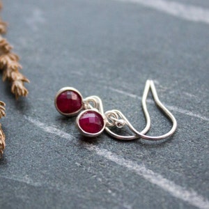 May include: Silver drop earrings featuring round, faceted, deep red gemstones. The earrings are presented on a dark grey surface, with a dried plant stem in the background. The gemstones are set in a silver bezel.