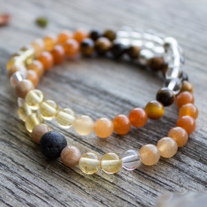 May include: Two beaded bracelets featuring a variety of gemstones in shades of yellow, orange, brown, and clear. One bracelet has a black lava bead. The bracelets are on a weathered wooden surface.