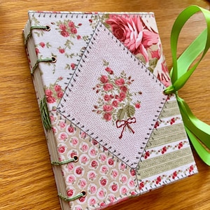 May include: A handmade journal with a patchwork cover featuring floral patterns in pink, red, and green. The journal is bound with green thread and has a green ribbon closure. The cover design includes a central embroidered floral motif.