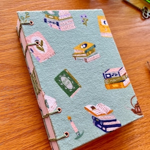 Peut inclure: Un carnet rectangulaire avec une couverture en tissu vert clair ornée d'un motif répétitif de livres empilés colorés, d'une bougie allumée et d'une théière. Le carnet est relié par une couture apparente en fil vert sur le côté gauche.