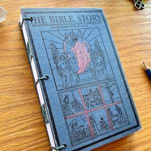 May include: A blue-covered book titled "THE BIBLE STORY" with religious illustrations and text. The cover art includes black and pink imagery. The book is bound with green thread and has white pages. The book's dimensions are not visible.