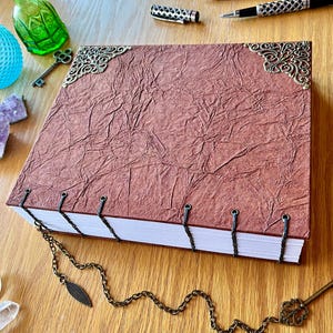 Puede incluir: Un diario hecho a mano con una cubierta texturizada de color marrón rojizo y esquinas decorativas de metal. El diario está encuadernado con hilo negro y presenta una cadena con un dije de hoja y una llave decorativa. Otros elementos incluyen una botella de vidrio verde y una esfera de vidrio azul.