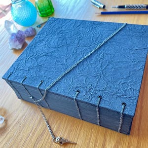 May include: A dark blue, textured journal with a silver chain closure and a decorative bird skull charm. The journal's pages are bound with silver thread. The cover has a crinkled paper texture. The journal is on a wooden surface.
