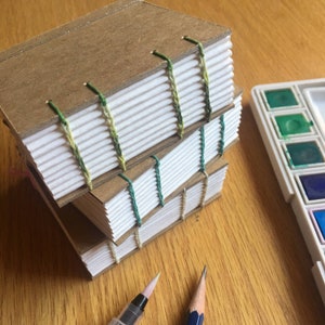 May include: Three handmade journals stacked on a wooden surface. The covers are brown, and the pages are white. The spines are stitched with green thread. A watercolor palette, a pencil, and a brush are also visible.