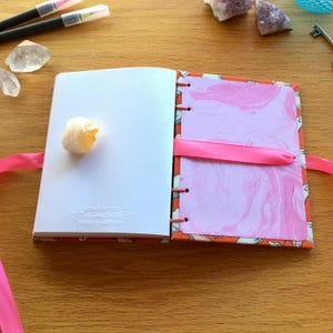May include: A pink and orange journal with a marbled pink paper insert. The journal has a pink ribbon bookmark and is closed. A white crystal is on the left page.