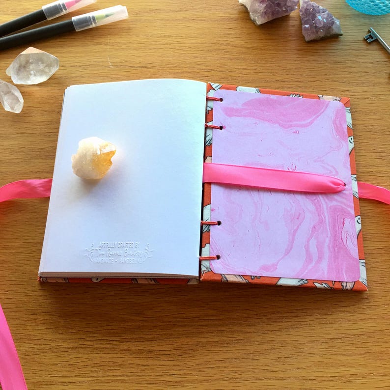 May include: A pink and orange journal with a marbled pink paper insert. The journal has a pink ribbon bookmark and is closed. A white crystal is on the left page.
