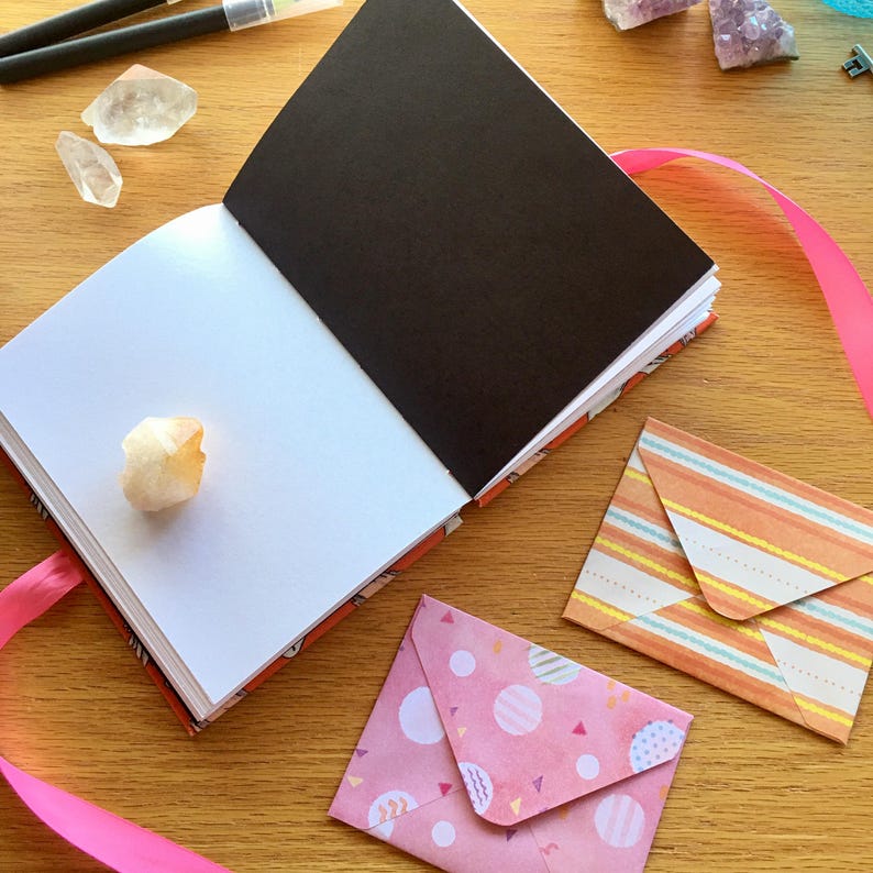 May include: An open blank journal with a black cover and a pink ribbon bookmark. The journal is on a wooden surface with two envelopes, one with a pink and white polka dot pattern and the other with a striped orange and white pattern.