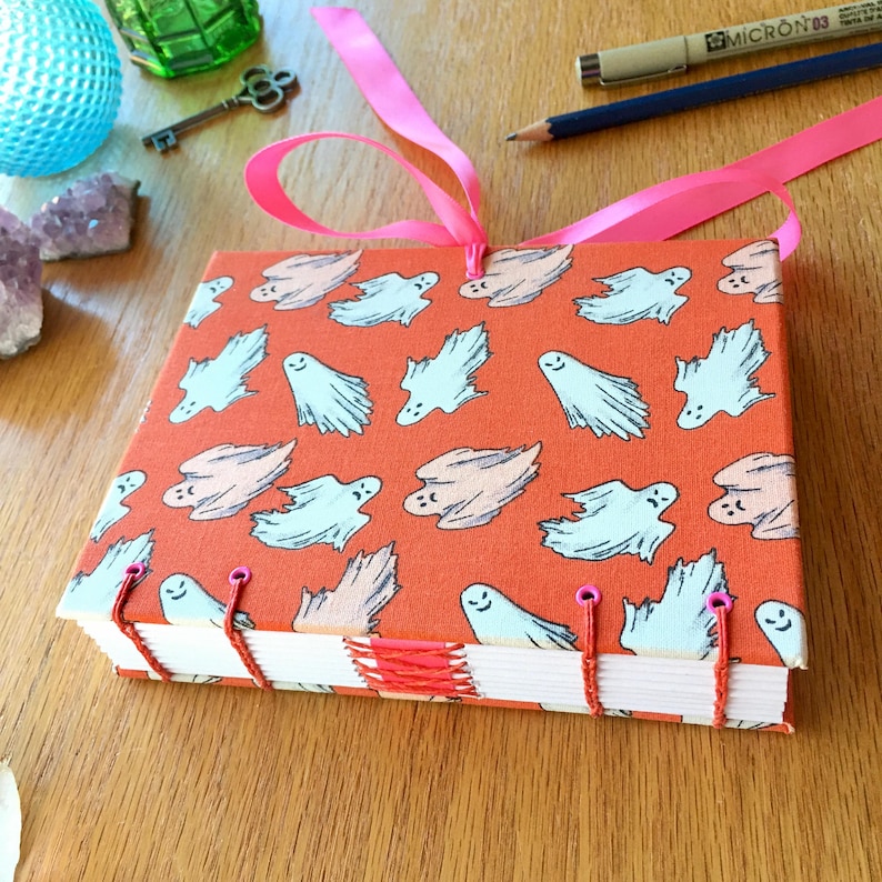 May include: A handmade journal with a bright orange cover featuring a repeating pattern of white ghosts with smiling faces. The journal is bound with red thread and has pink ribbon ties.
