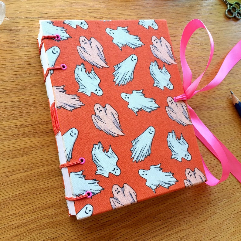 May include: A handmade orange journal with a ghost pattern. The journal has pink ribbon ties and is closed. The journal has pink stitching and eyelets.