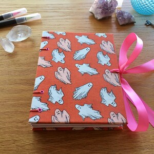 May include: A handmade orange journal with a ghost pattern and a pink ribbon closure. The journal has a stitched binding and is perfect for writing or drawing.