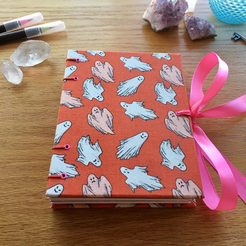 May include: A handmade orange journal with a ghost pattern and a pink ribbon closure. The journal has a stitched binding and is perfect for writing or drawing.