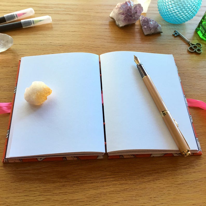 May include: An open notebook with a white, blank page on each side. A wooden fountain pen is lying on the right page. A small, yellow crystal is on the left page. The notebook has a red and white patterned cover.