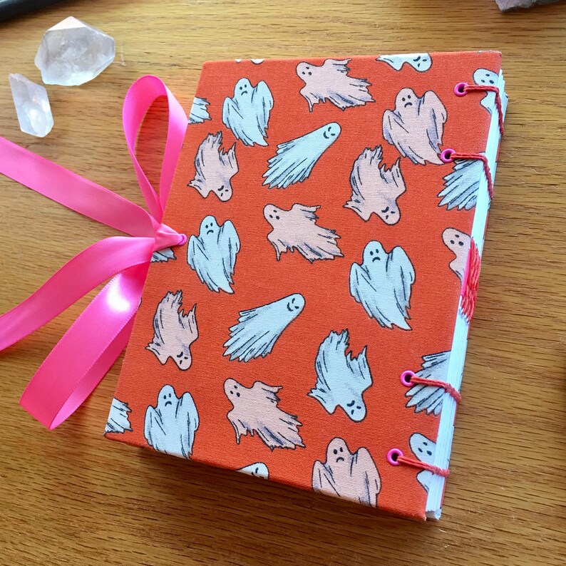 May include: A handmade journal with a bright orange cover featuring a repeating pattern of white and blue ghosts. The journal is bound with pink ribbon and has pink stitching along the spine.