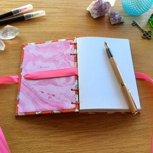 May include: An open notebook with a pink marbled cover and a pink ribbon bookmark. The notebook has blank pages and a gold pen is lying on the open page.
