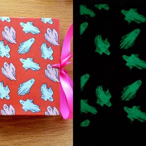 May include: A small orange book with a pink ribbon closure. The cover is decorated with a repeating pattern of white ghosts. The book glows in the dark, revealing green ghosts.